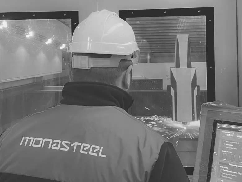 Mona Steel operator in a hard hat running a fiber laser cutting machine, sparks at the cutting head